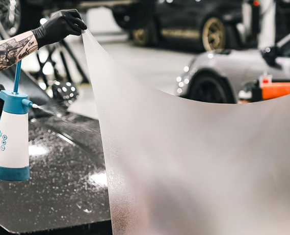 Two workers in a car shop stretch a transparent protective film over the hood of a dark luxury sports car.