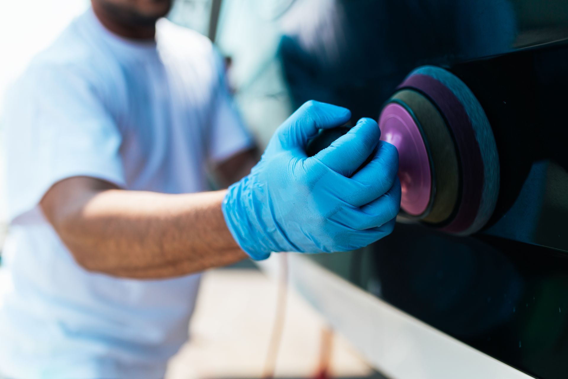 A person wearing blue gloves polishes a car’s dark, glossy exterior with a multi-layered buffing pad.