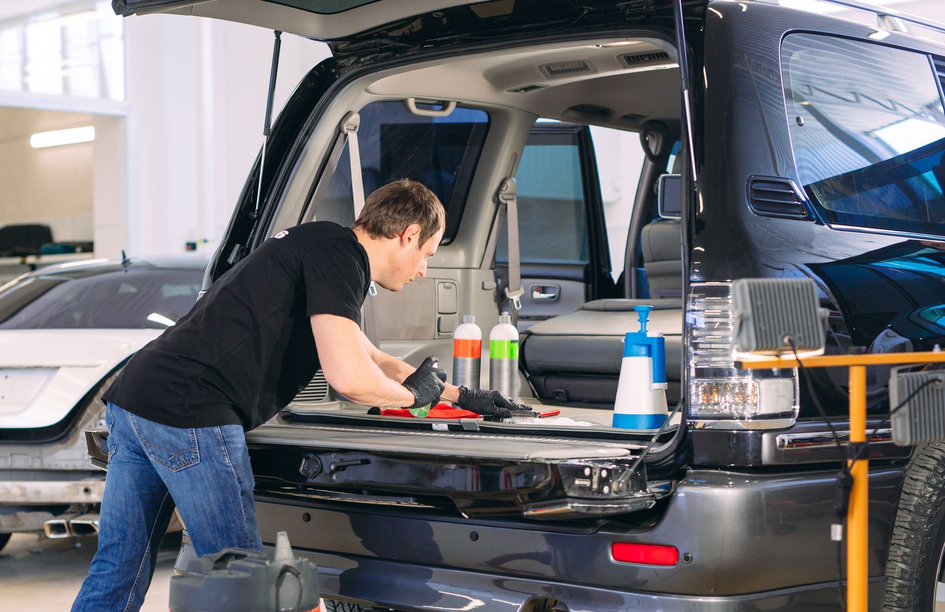 A person in a black shirt and jeans cleans the trunk of a black SUV inside an auto detailing shop.