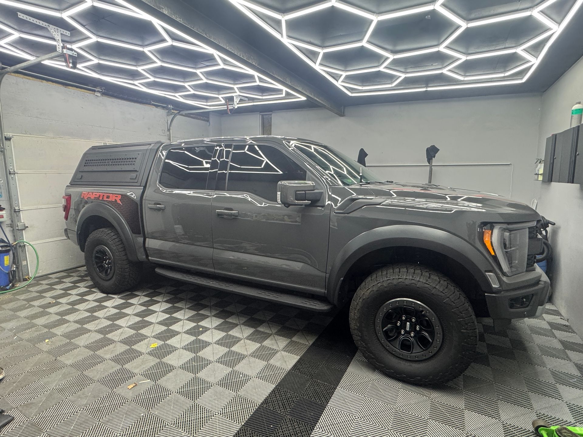A close-up of a black off-road vehicle wheel with a simulated beadlock rim design and textured all-terrain tire.