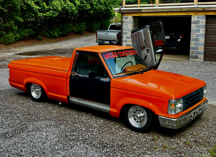 A modified bright orange pickup truck with an open gull-wing driver's side door parked on a gravel driveway.