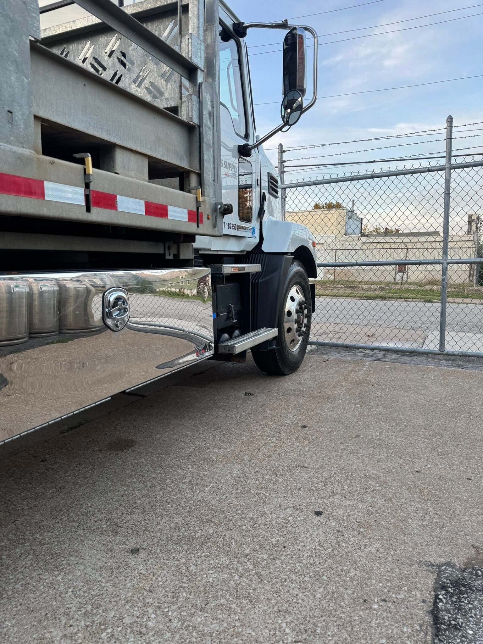 A white flatbed truck parked on a gravel lot next to a chain-link fence with barbed wire.