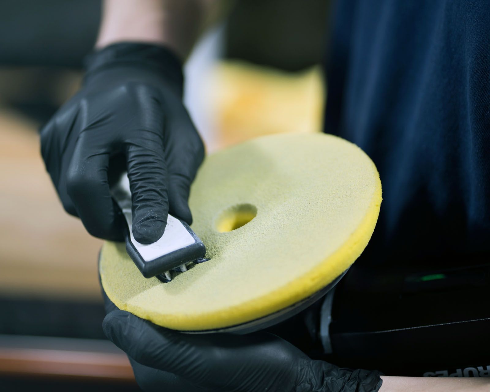 A person in black gloves cleans a yellow foam polishing pad with a specialized cleaning brush.