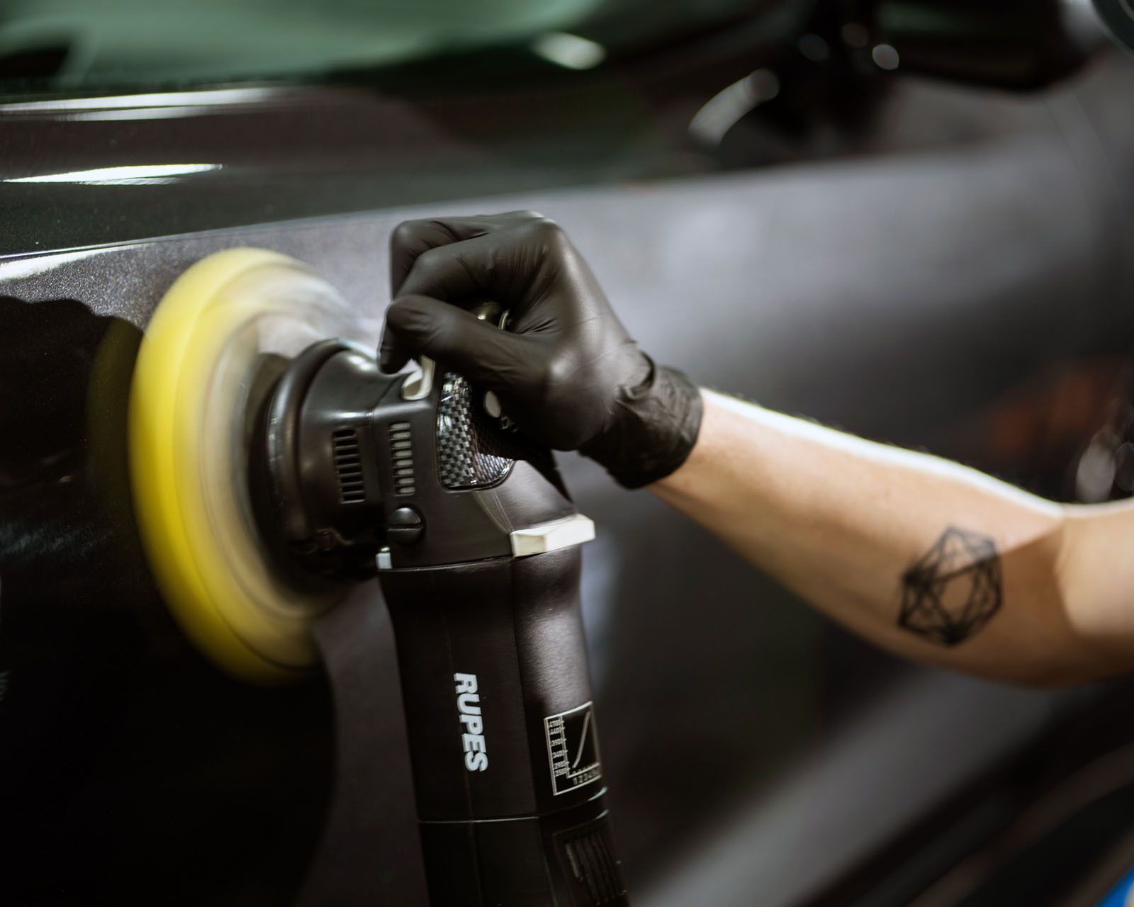 A person wearing black gloves uses a yellow-padded RUPES polisher on a dark car surface.