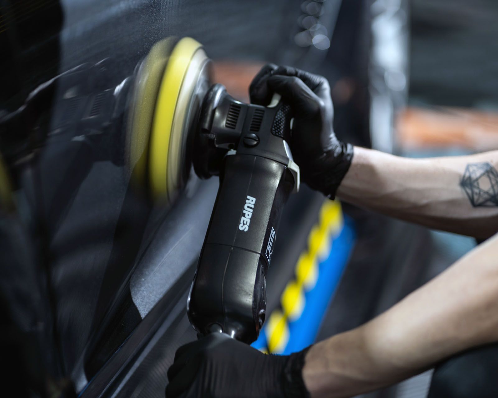A person wearing black gloves uses a black Rupes dual-action polisher with a yellow foam pad on a dark car surface.