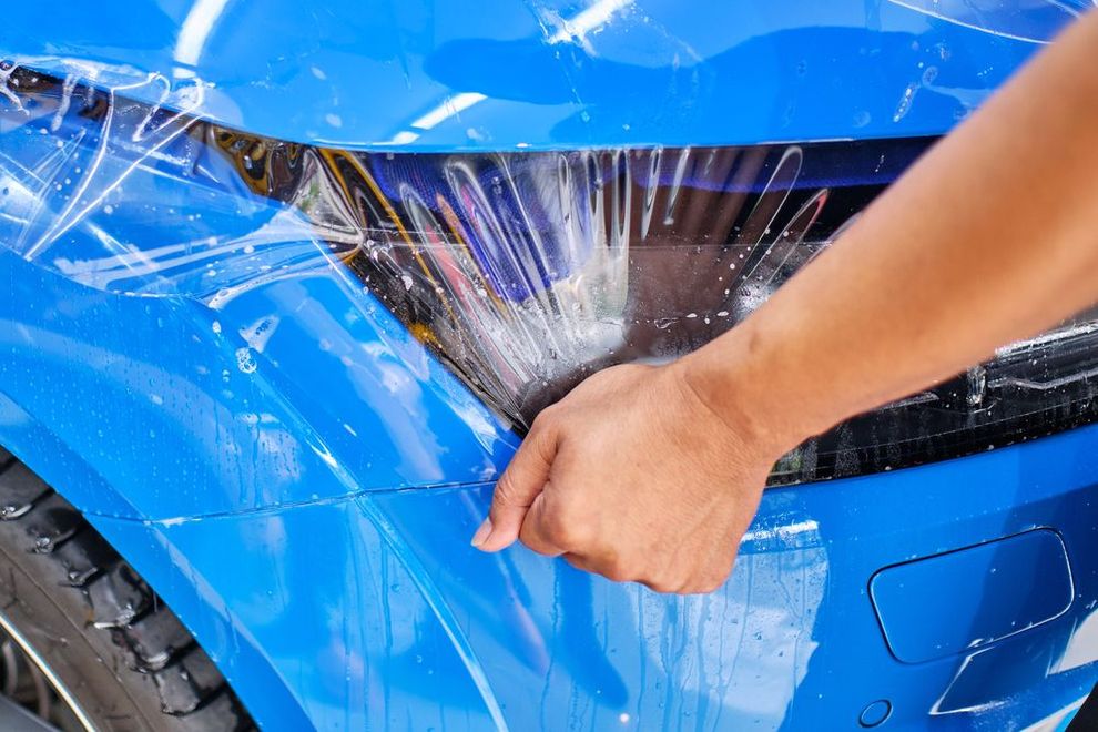 A hand applies a protective clear film to the headlight of a bright blue car.