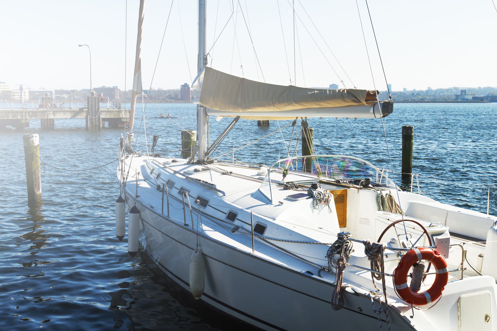 A white sailboat docked at a pier on a bright, sunny day with a city skyline visible in the distance.