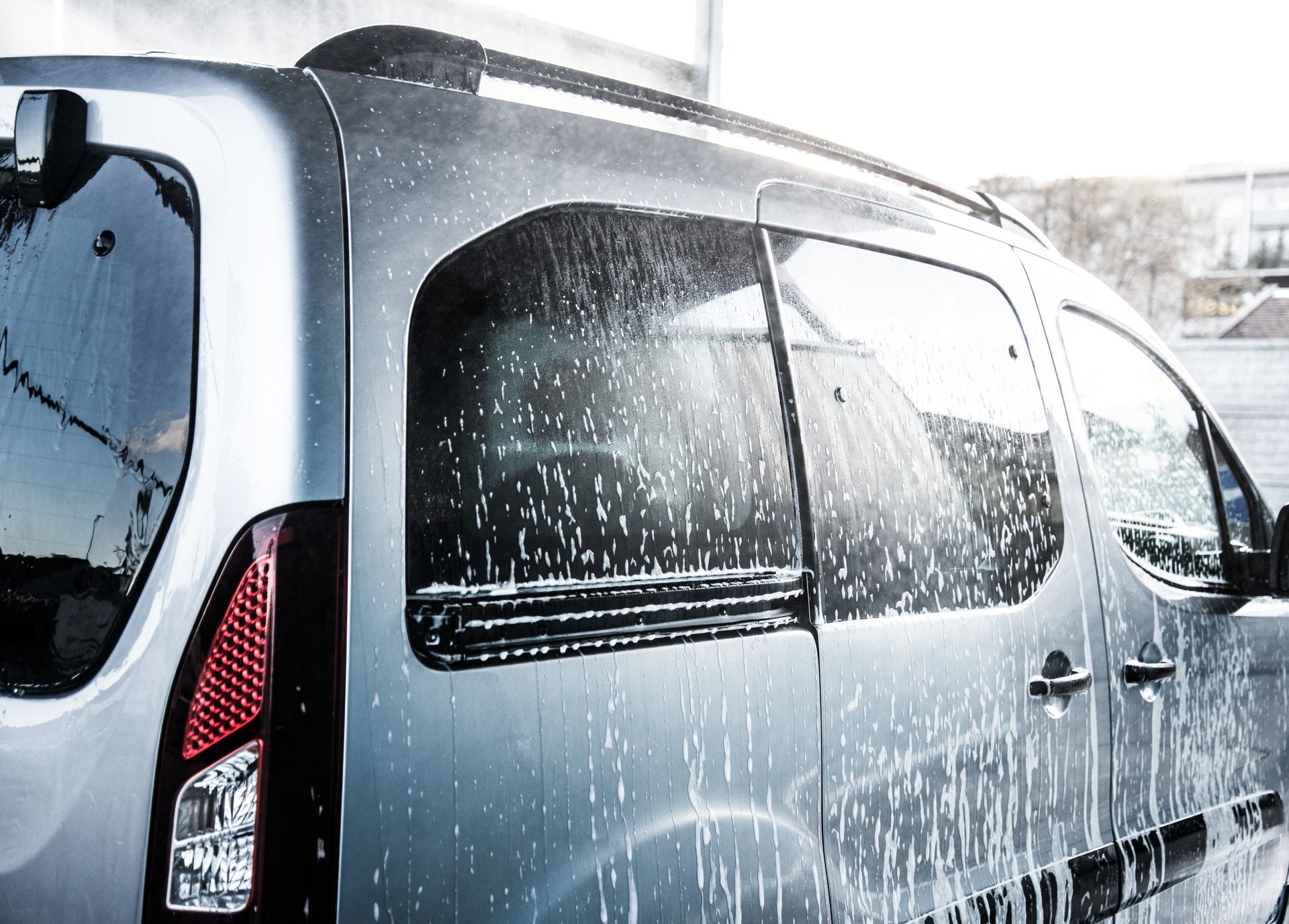 A silver minivan being washed, covered in soap suds and water streaks.