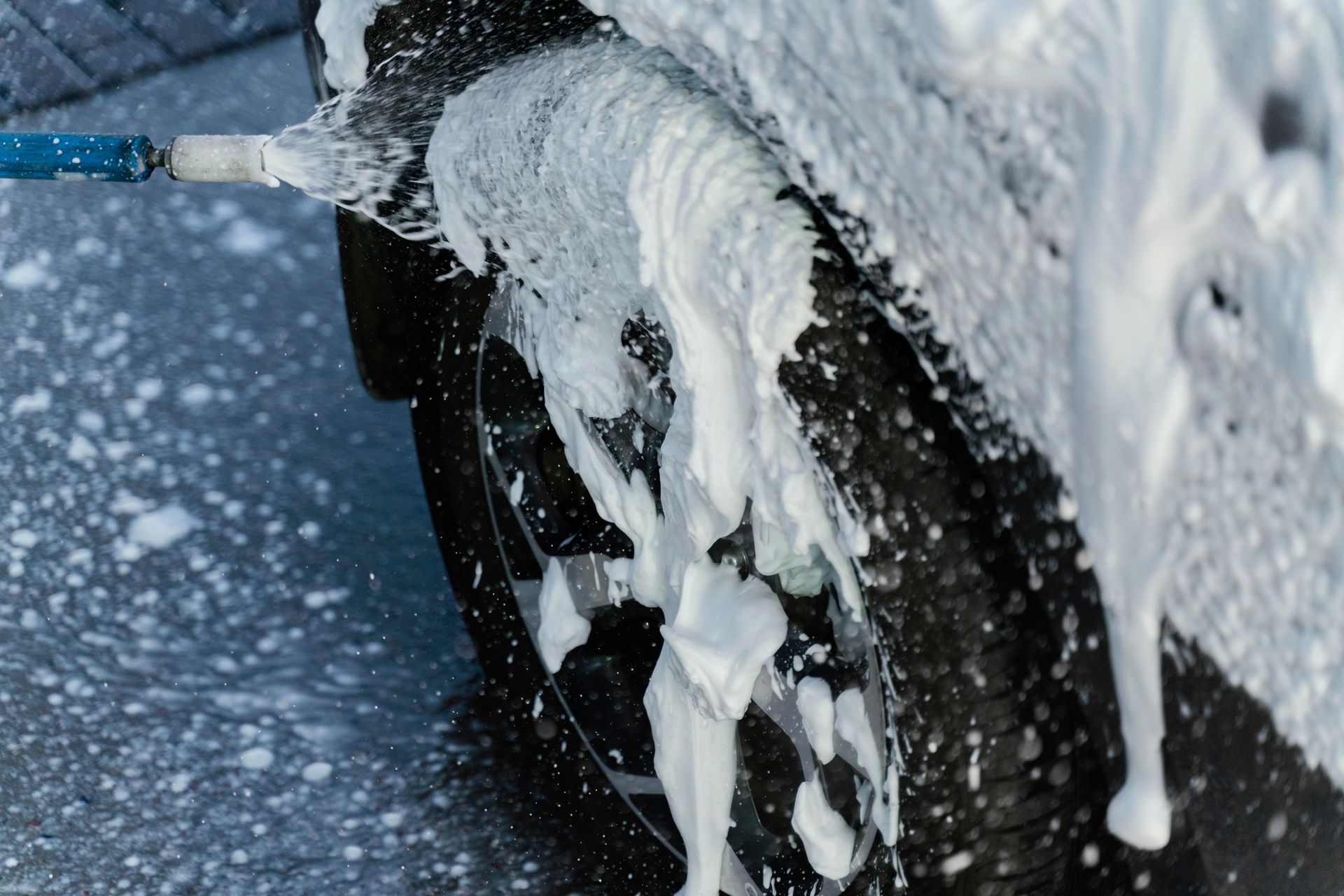 A close-up of a blue hose spraying thick white cleaning foam onto a dark car tire and wheel.