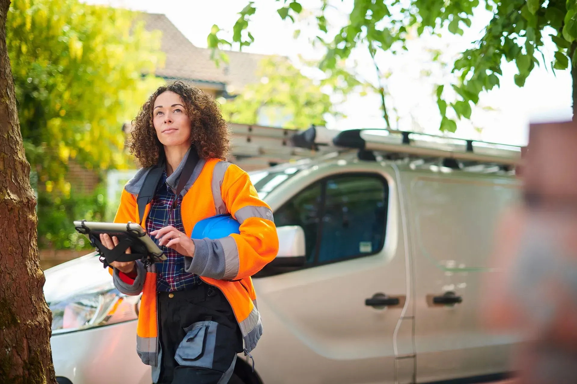 Woman in orange safety vest holding tablet and blue helmet next to work van.