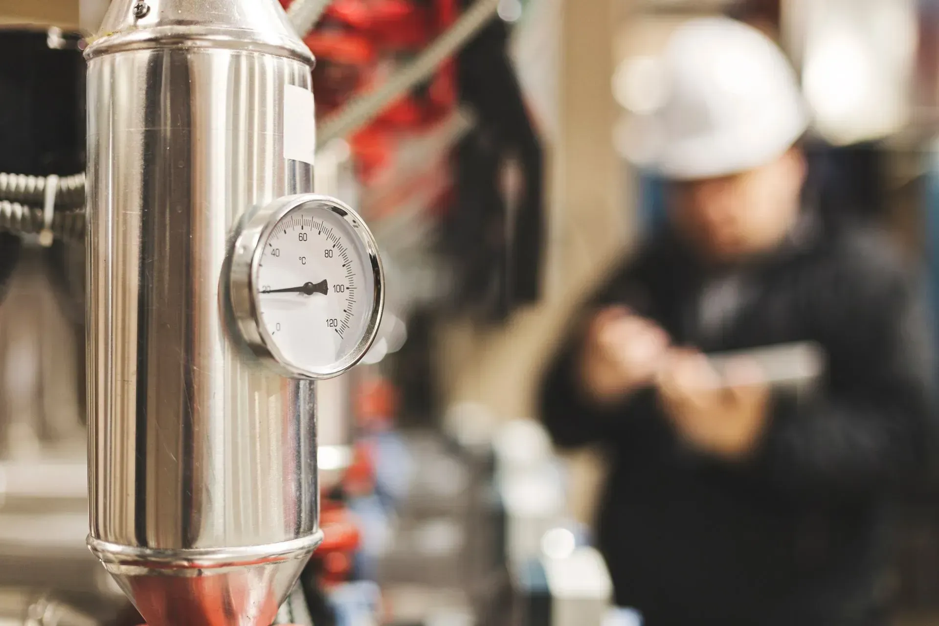 Pressure gauge on stainless steel pipe; worker in hard hat and dark clothing in background.