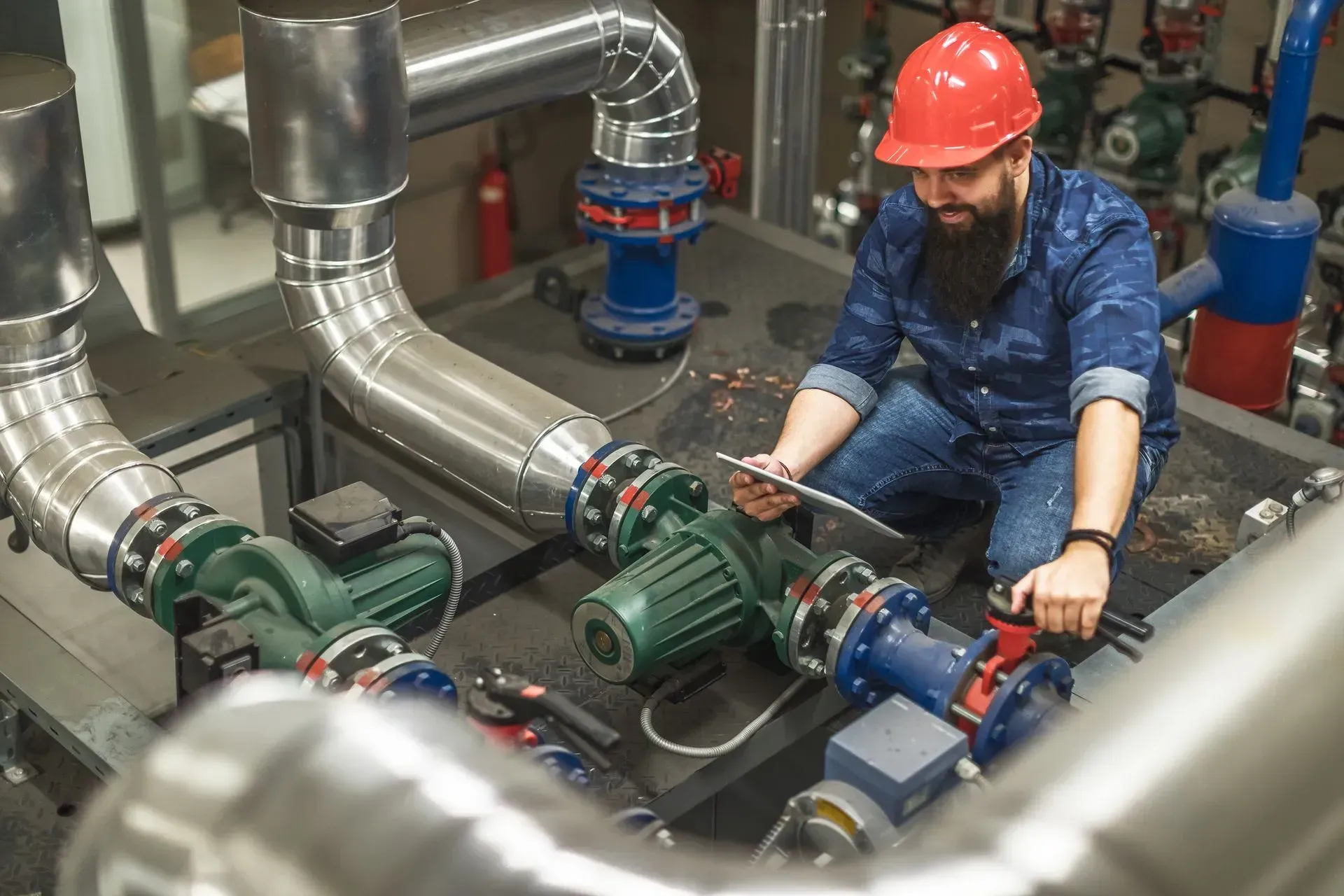 Engineer in red hard hat inspecting industrial pipes and machinery with a tablet.