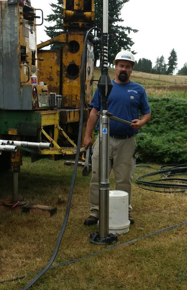 Man in blue shirt, hard hat, holding a submersible water pump outdoors near drilling equipment. Man in blue shirt, hard hat, holding a submersible water pump outdoors near drilling equipment.