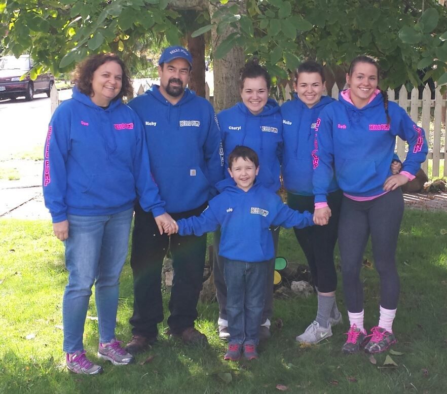 Six people in blue hoodies holding hands in front of a tree and white fence.
