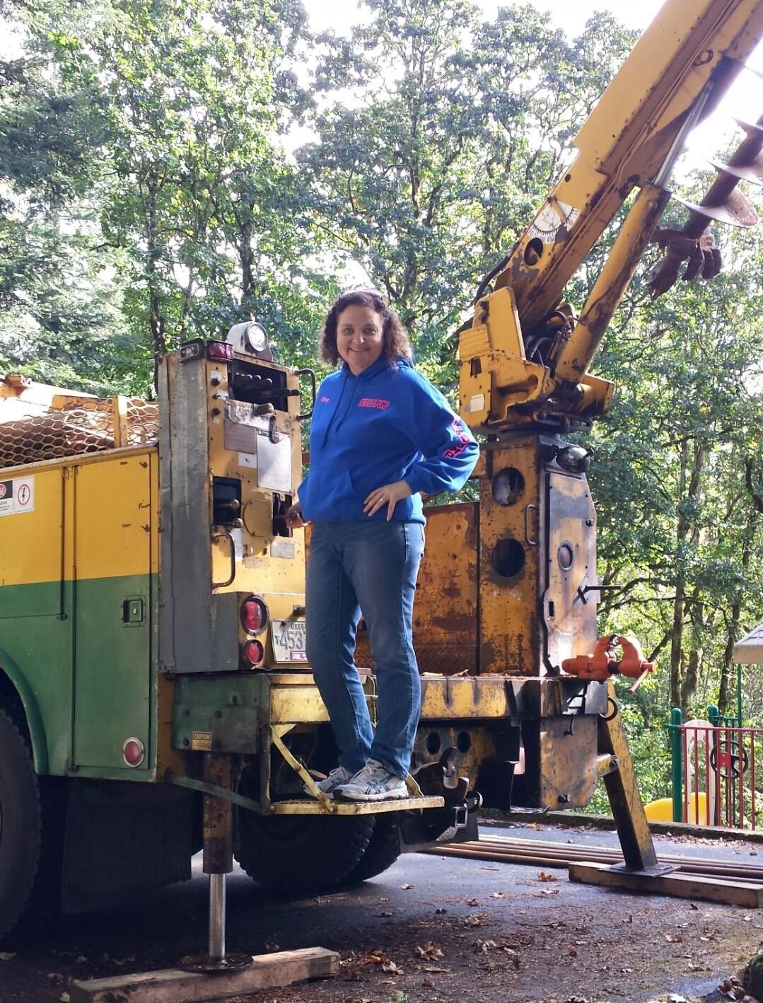Woman in blue hoodie standing on a large yellow truck with a raised crane, trees in background. Woman in blue hoodie standing on a large yellow truck with a raised crane, trees in background.