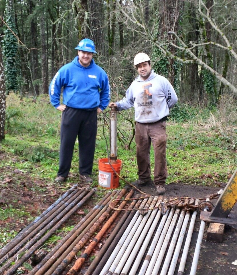Two workers stand by well drilling equipment outdoors; one holds a tool. Two workers stand by well drilling equipment outdoors; one holds a tool.