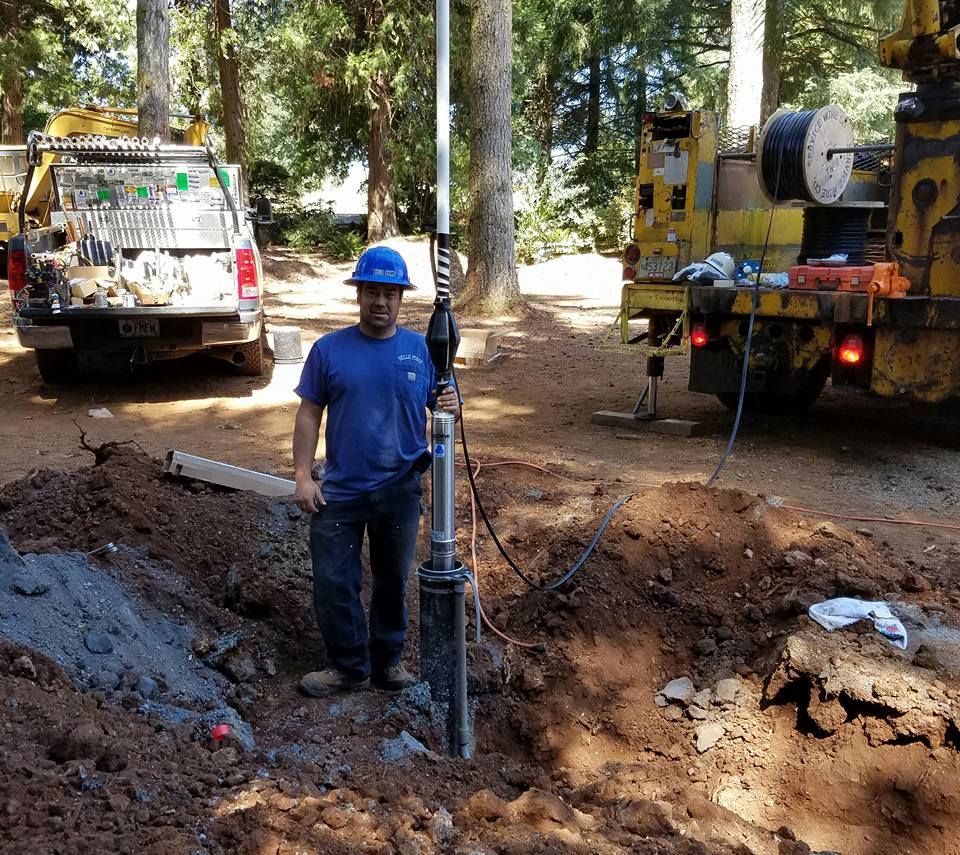 Man in blue shirt and hard hat holding well equipment near trucks and excavation.