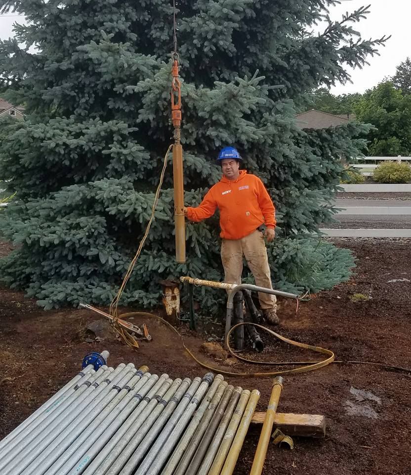 Man in orange shirt by drilling equipment near a blue spruce. Brown dirt and pipes visible. Man in orange shirt by drilling equipment near a blue spruce. Brown dirt and pipes visible.