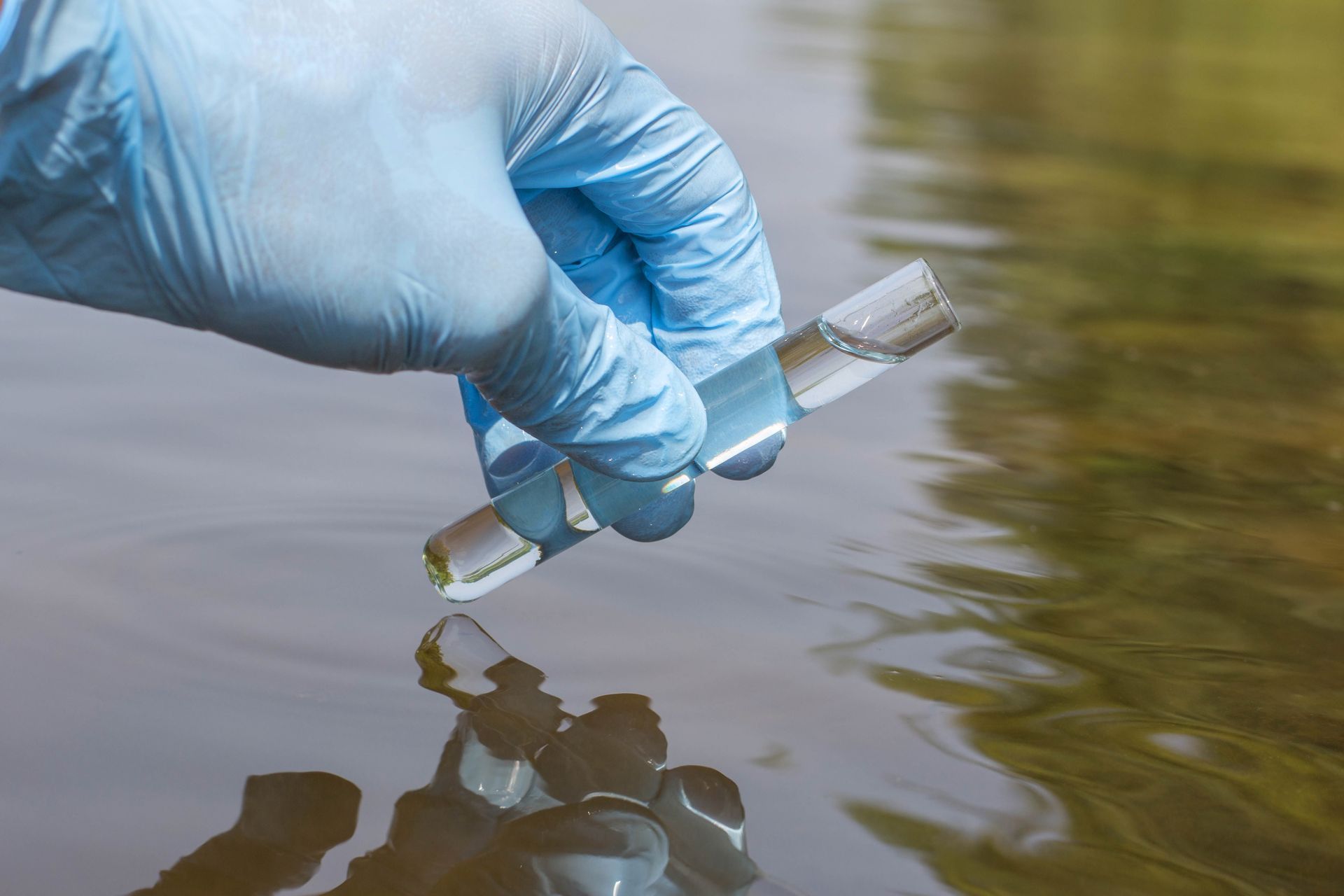 Gloved hand collecting a water sample in a vial from a body of water outdoors.