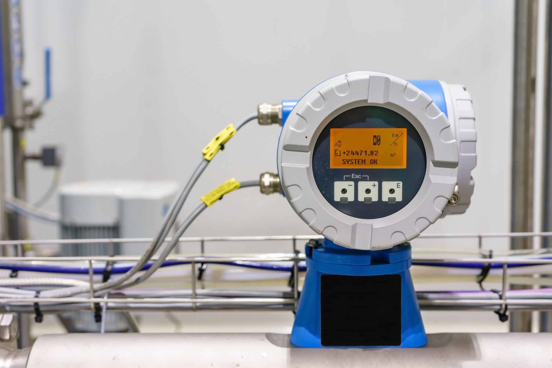 Industrial flow meter with a digital display and connected wiring, mounted on a stainless steel pipe. Industrial flow meter with a digital display and connected wiring, mounted on a stainless steel pipe.