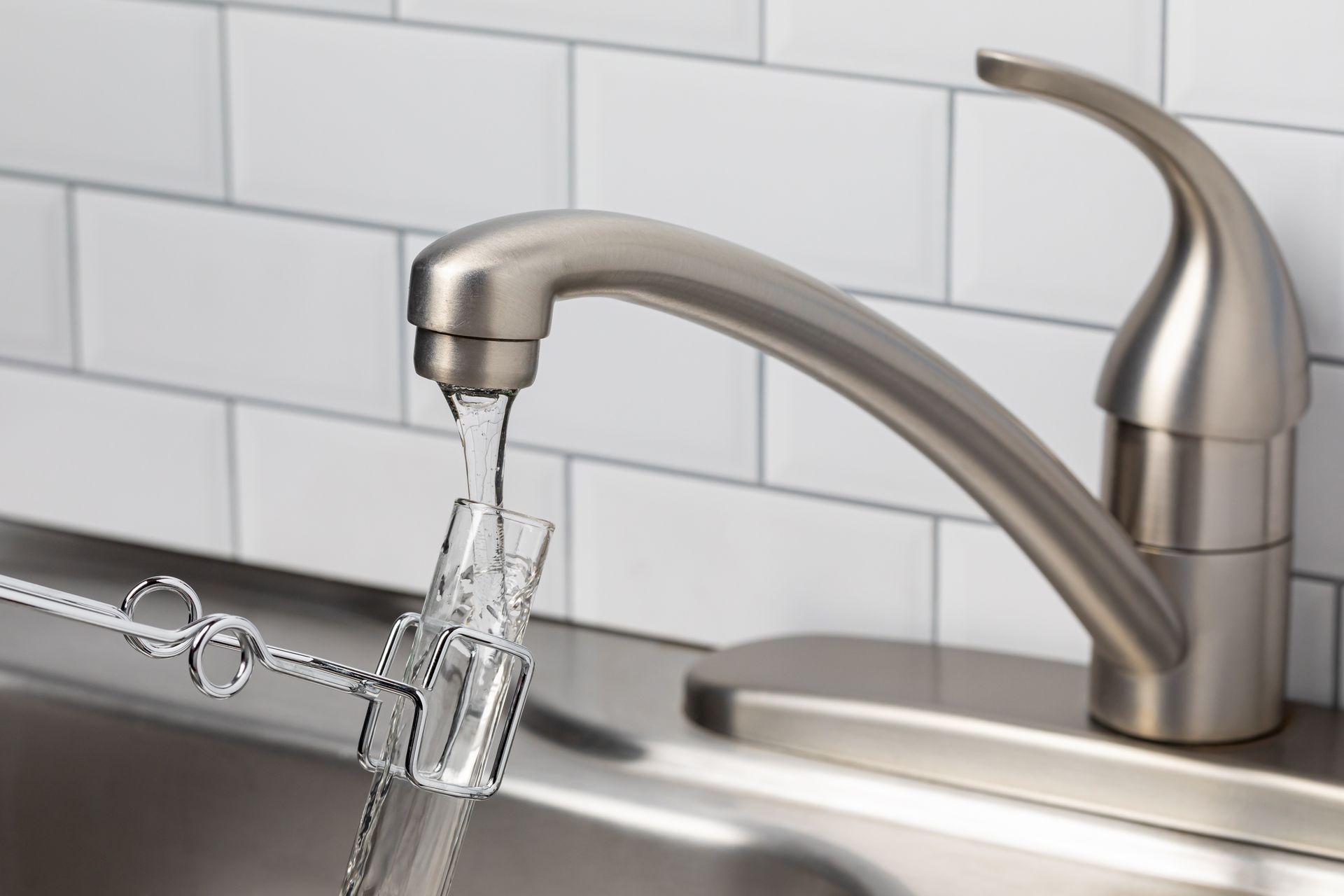 Faucet dispensing water into a small glass beaker held in a metal clamp, over a stainless steel sink.