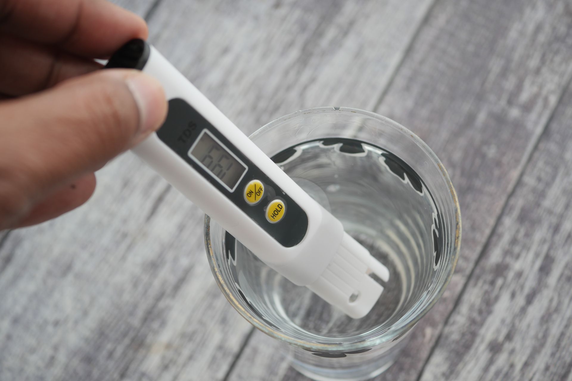 Hand holding a water quality tester in a glass of water on a wooden surface.