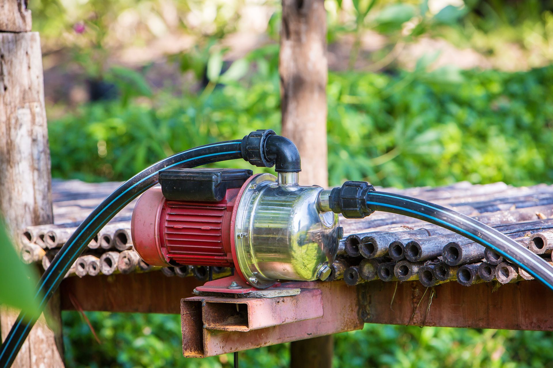 Red and silver water pump connected to black hoses, sitting on a wooden structure outdoors.