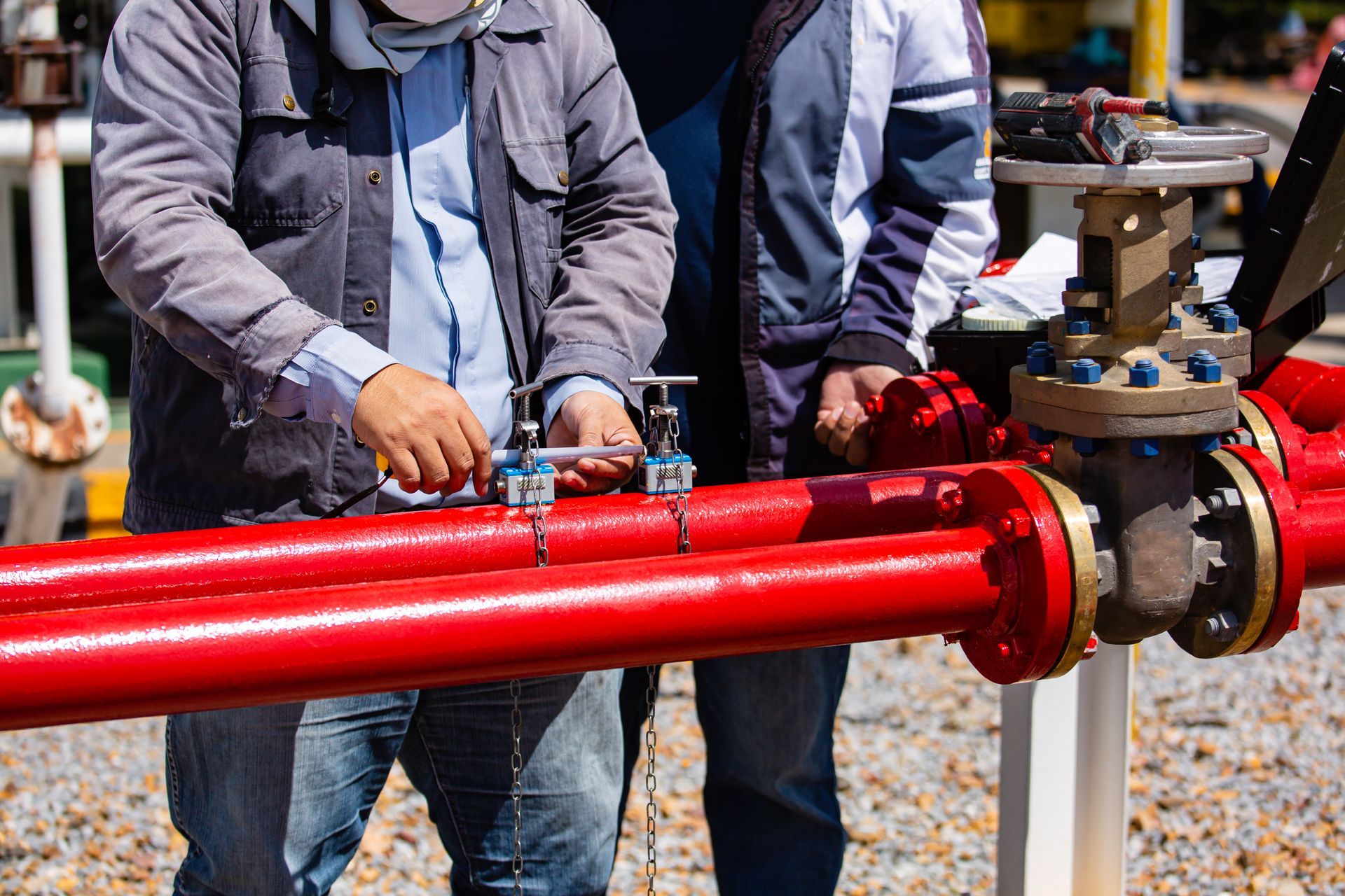 Two workers inspect red pipes and a valve at an industrial site. Two workers inspect red pipes and a valve at an industrial site.