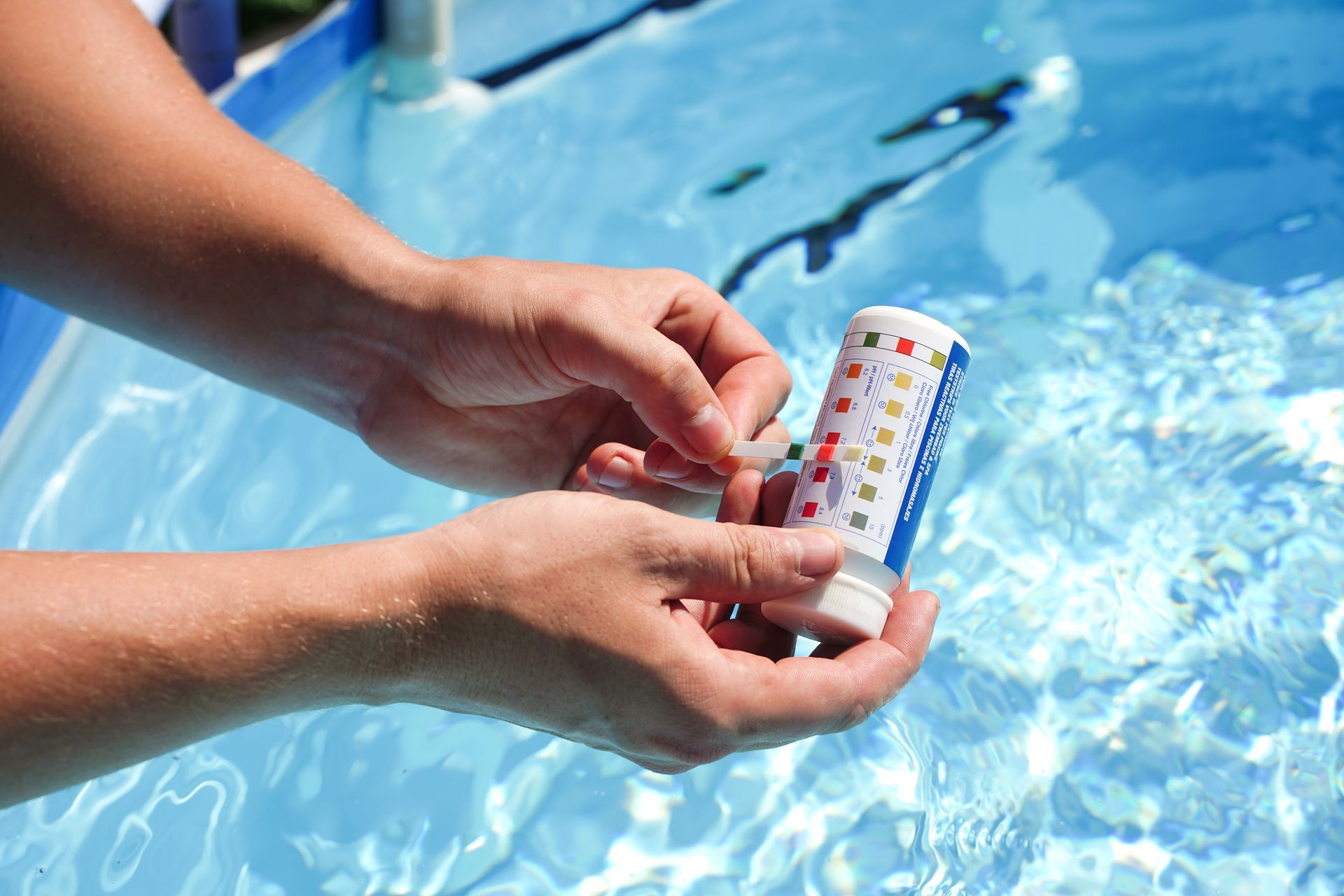 Hands holding a pool water testing kit over a blue pool, comparing color results.