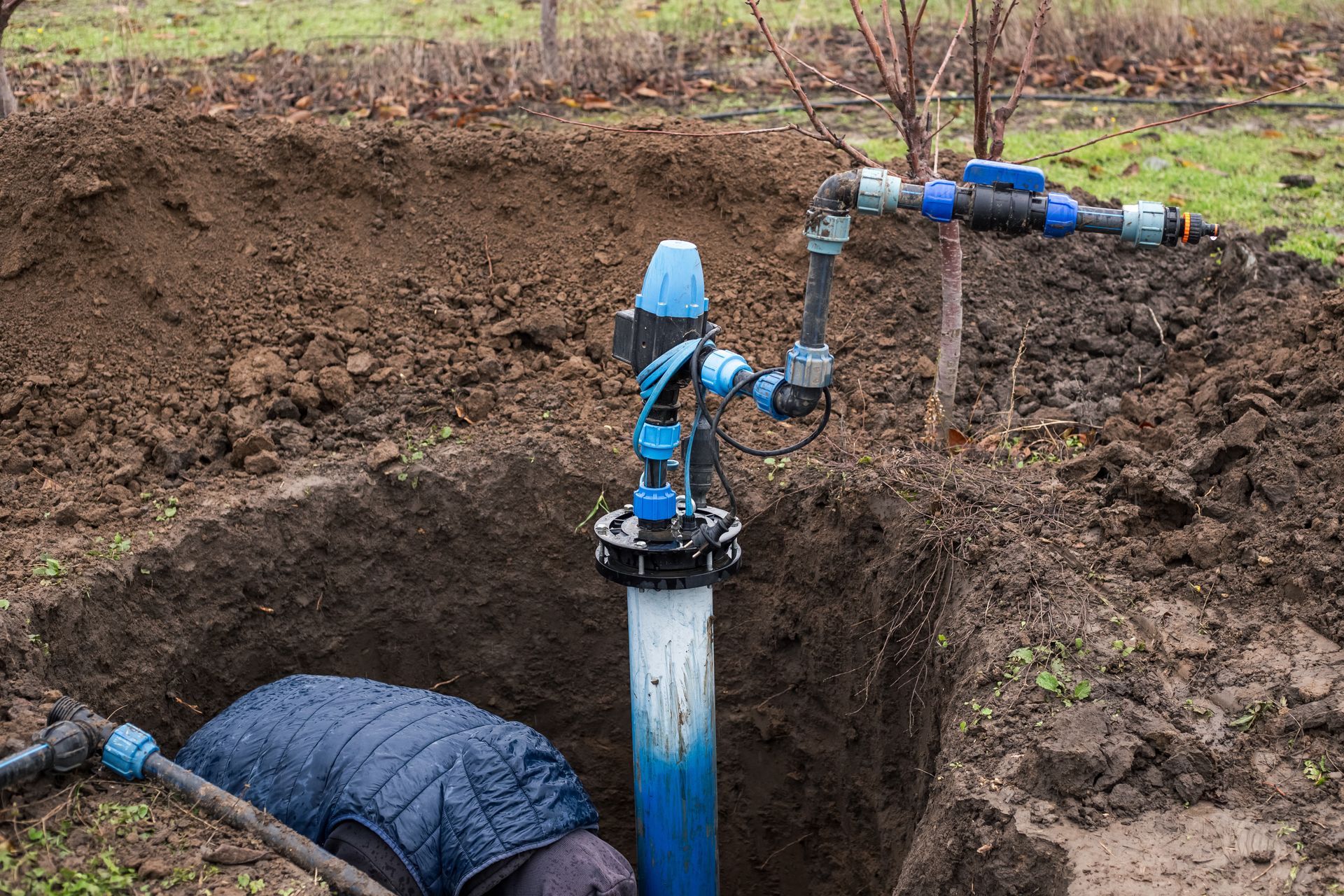Person working on water well in a hole in the ground, with blue pipes and valves visible. Person working on water well in a hole in the ground, with blue pipes and valves visible.