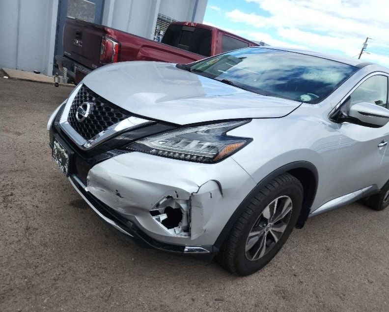 A silver nissan murano with a damaged front bumper is parked in front of a building.