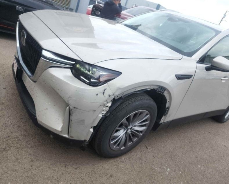 A white car with a damaged front bumper is parked in a parking lot.