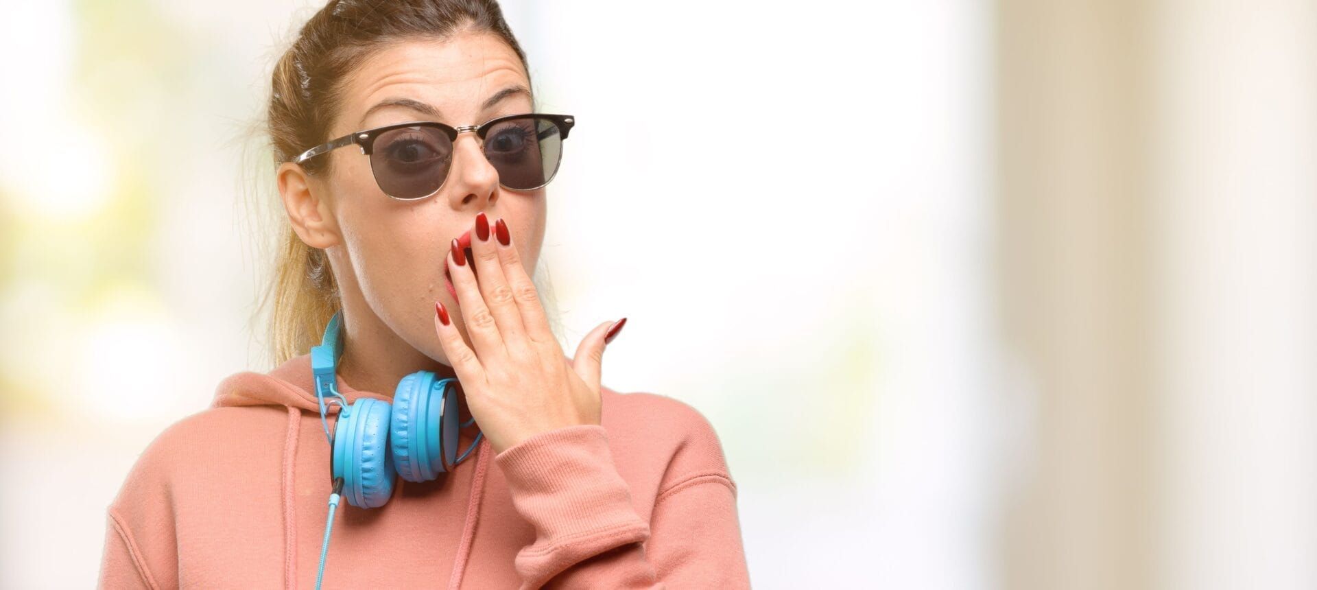 A young woman wearing headphones and sunglasses is yawning and covering her mouth with her hand.