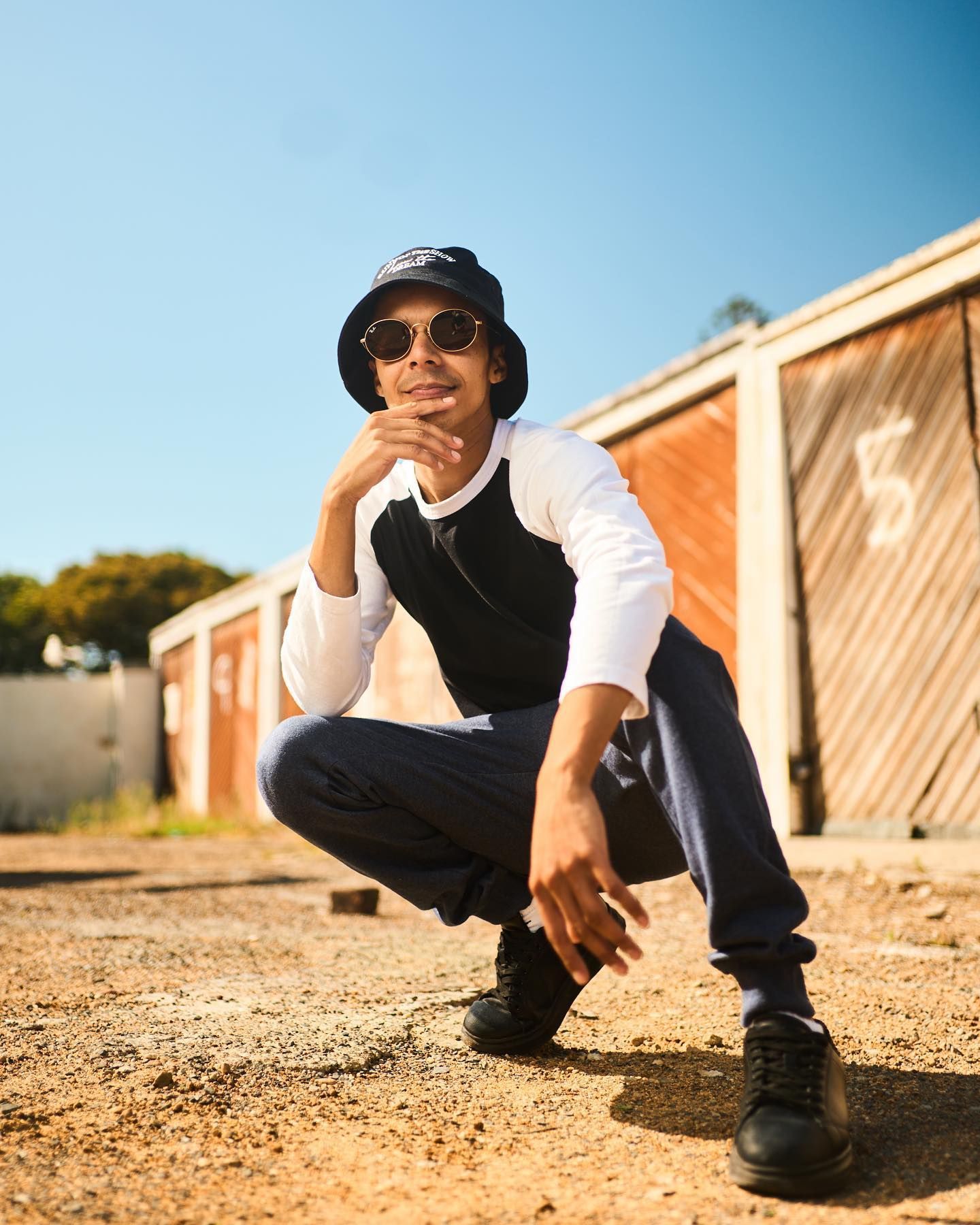 A man wearing a hat and sunglasses is squatting down on the ground.