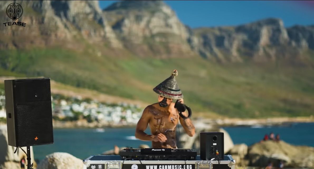 A man is playing a dj set on a beach with mountains in the background.