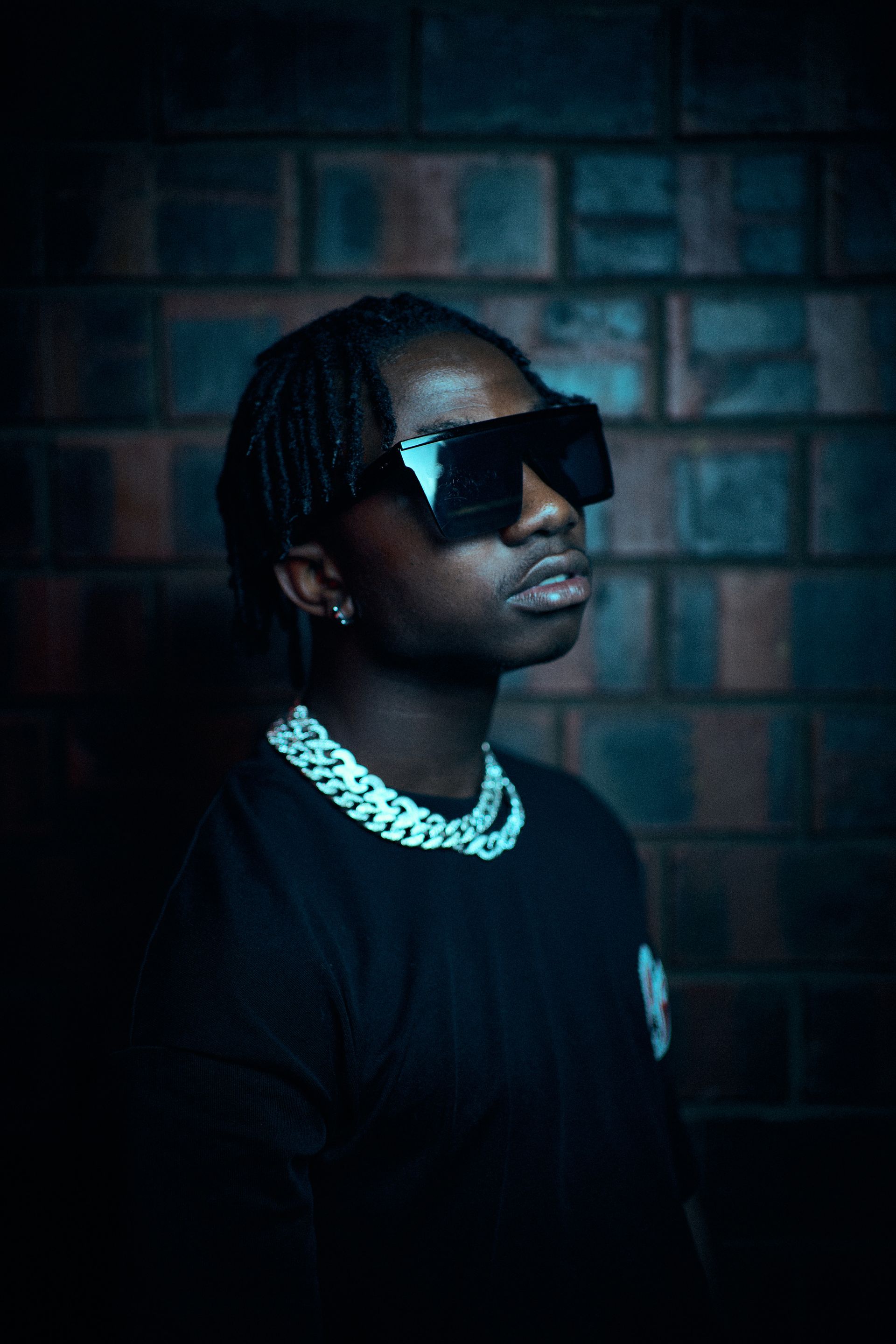 A man wearing sunglasses and a necklace is standing in front of a brick wall.