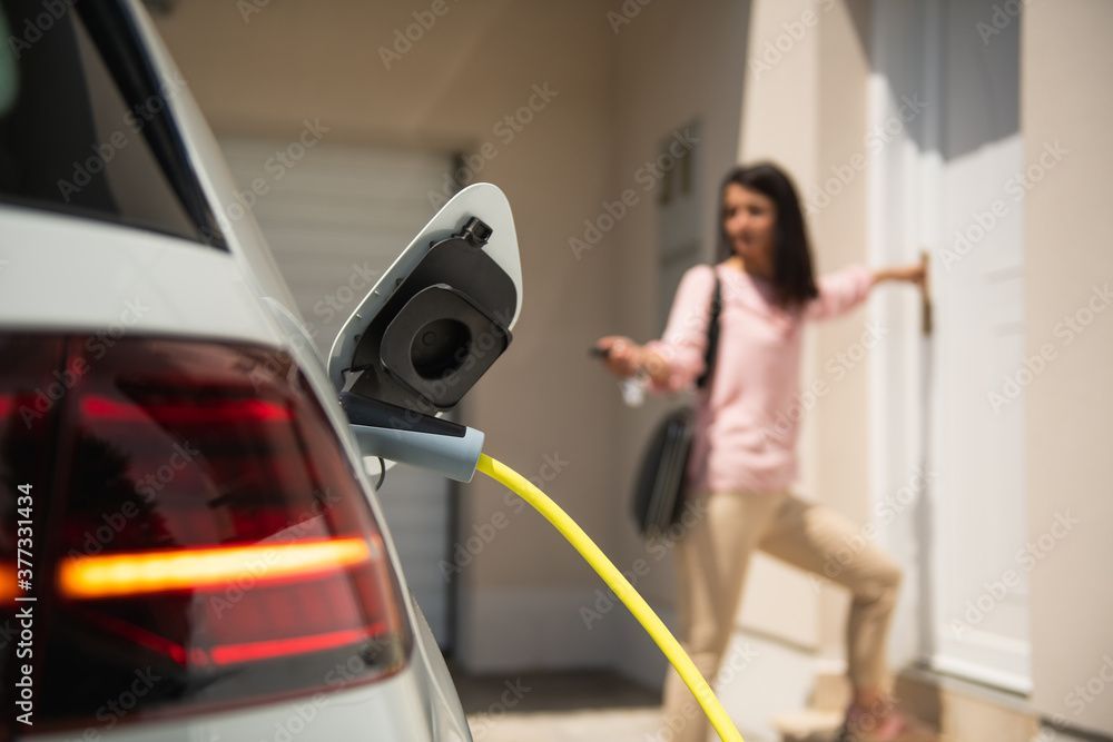 Une femme se tient près d'une maison, tenant un porte-clés de voiture. Une voiture électrique est branchée avec un câble de recharge jaune.