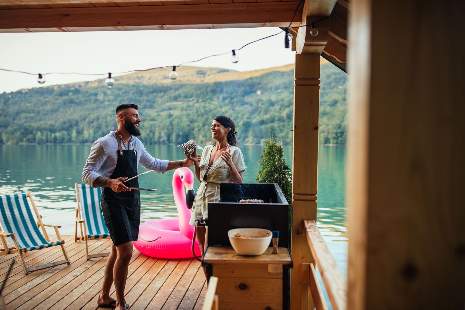 A man and a woman are having a barbecue on a dock next to a lake.