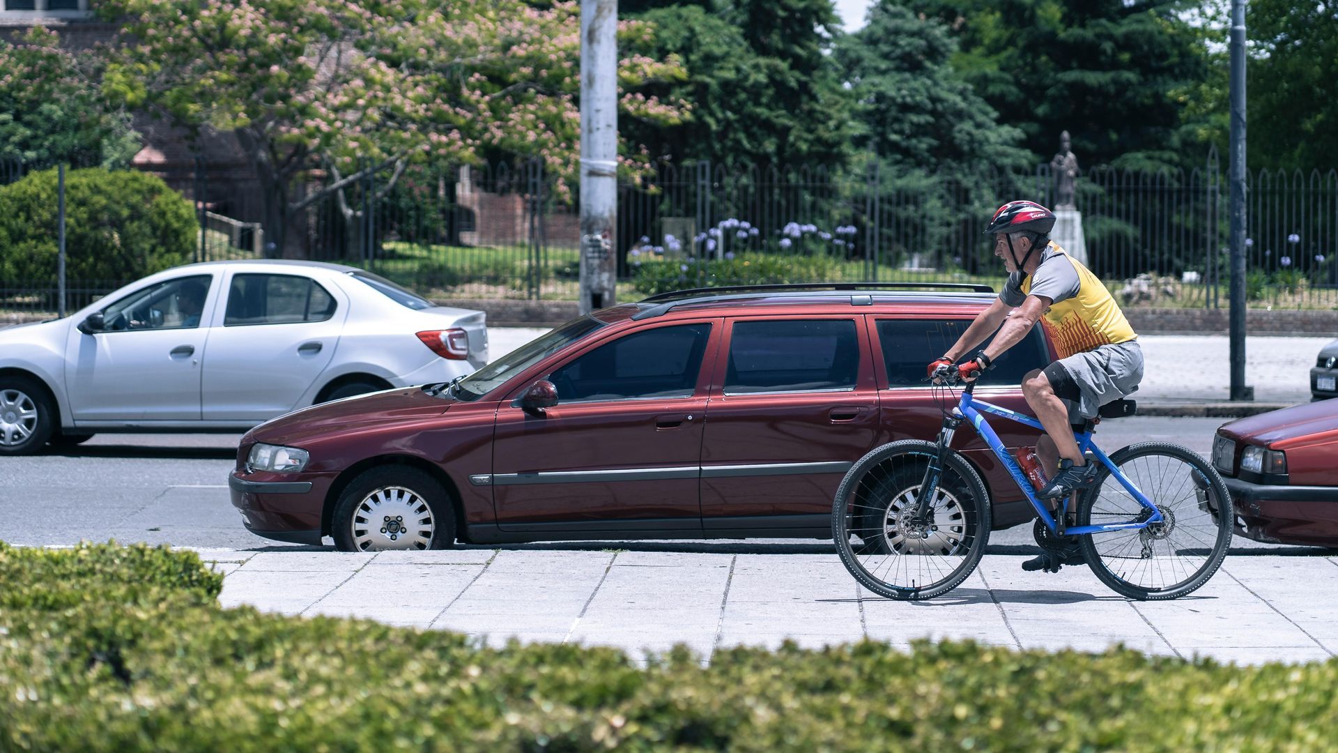 A cyclist in a yellow jersey rides a blue bike on a city sidewalk past a row of parked cars on a sunny day.