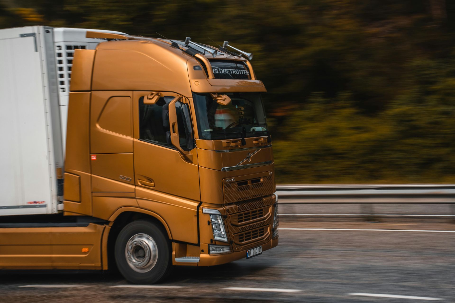 A metallic gold Volvo semi-truck driving on a highway with a blurred background of trees.