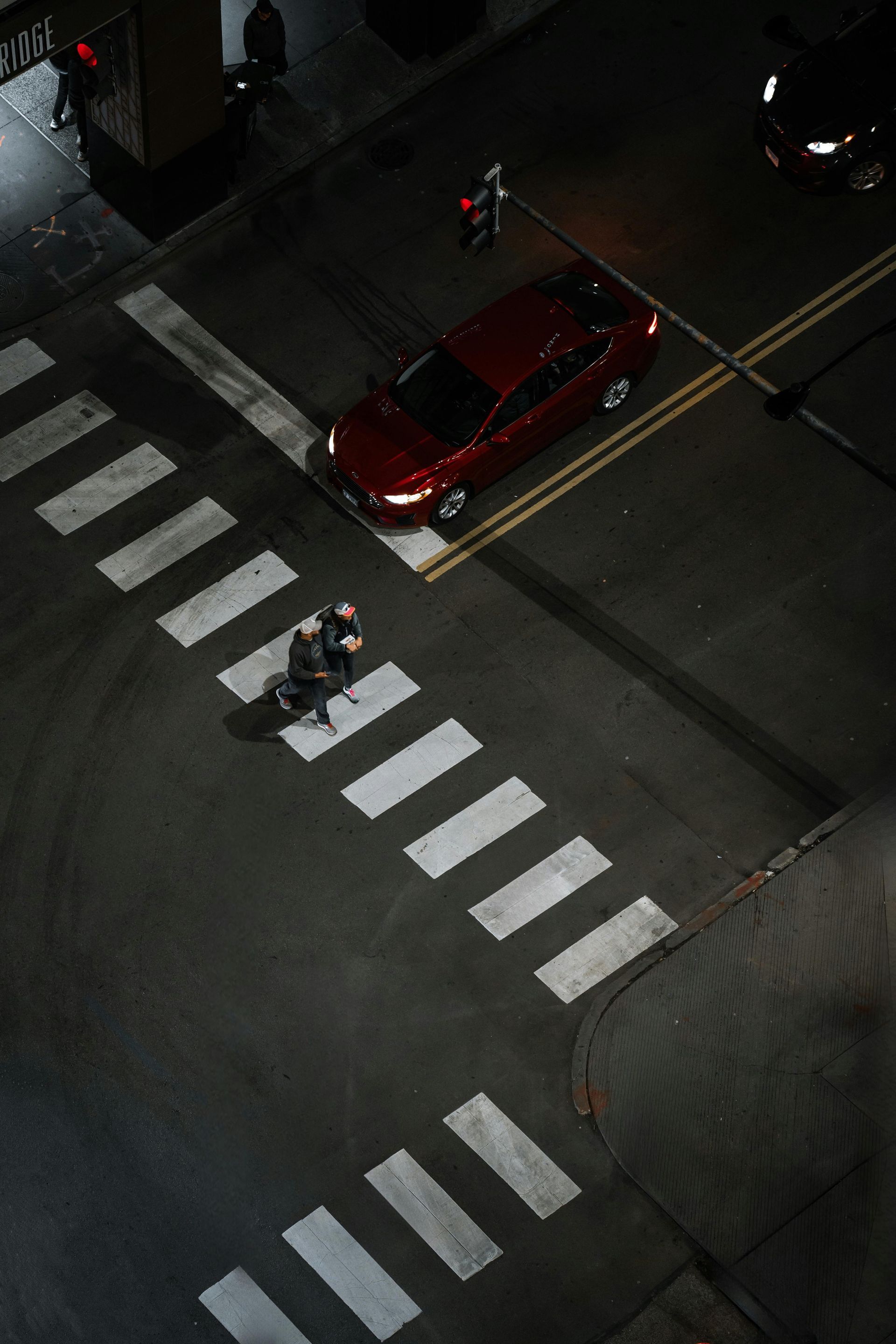 An aerial view of a person crossing a dark city street on a white-striped crosswalk as a red car waits nearby.