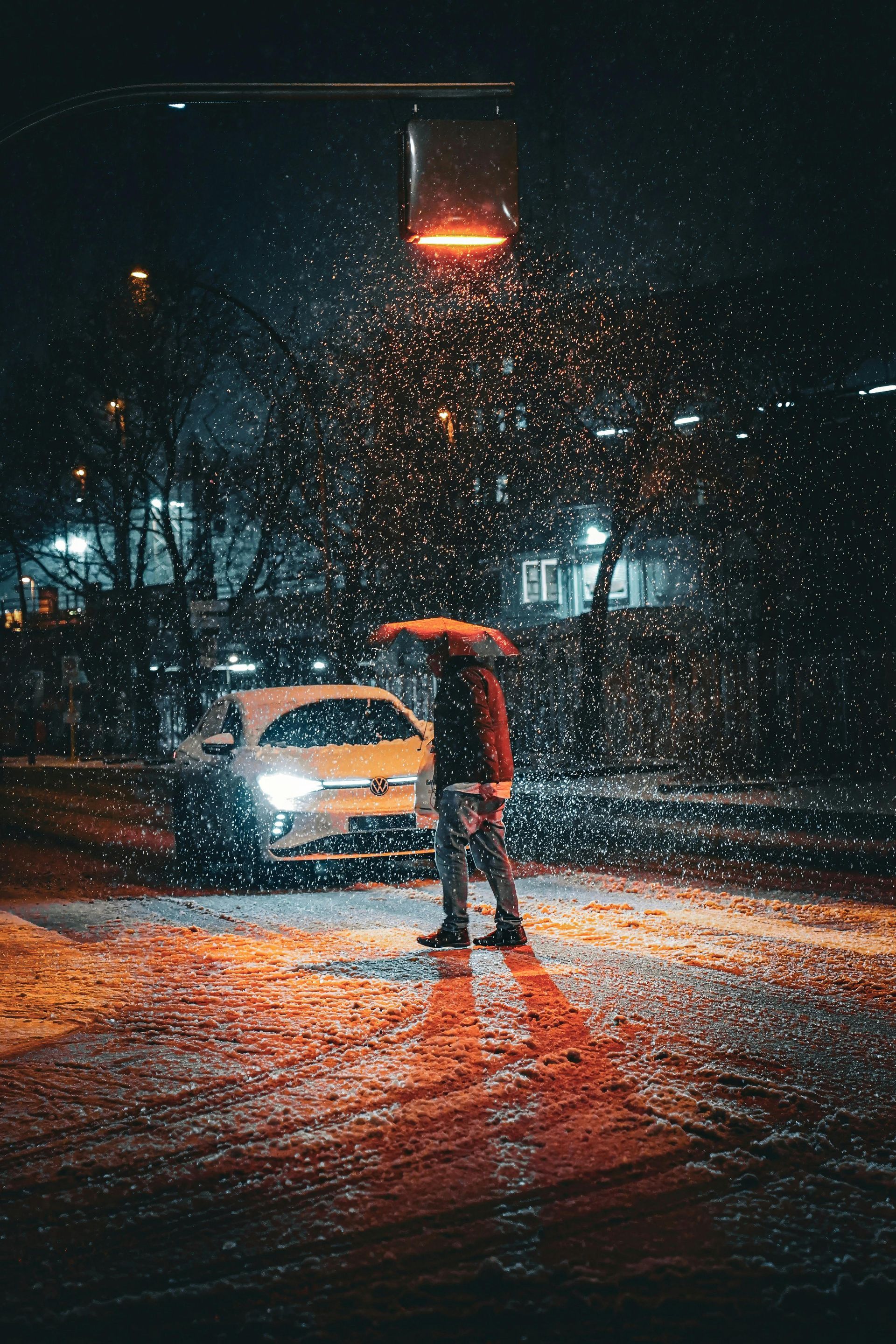 A person stands under a streetlight at night during a snowfall, with a parked car visible in the background.