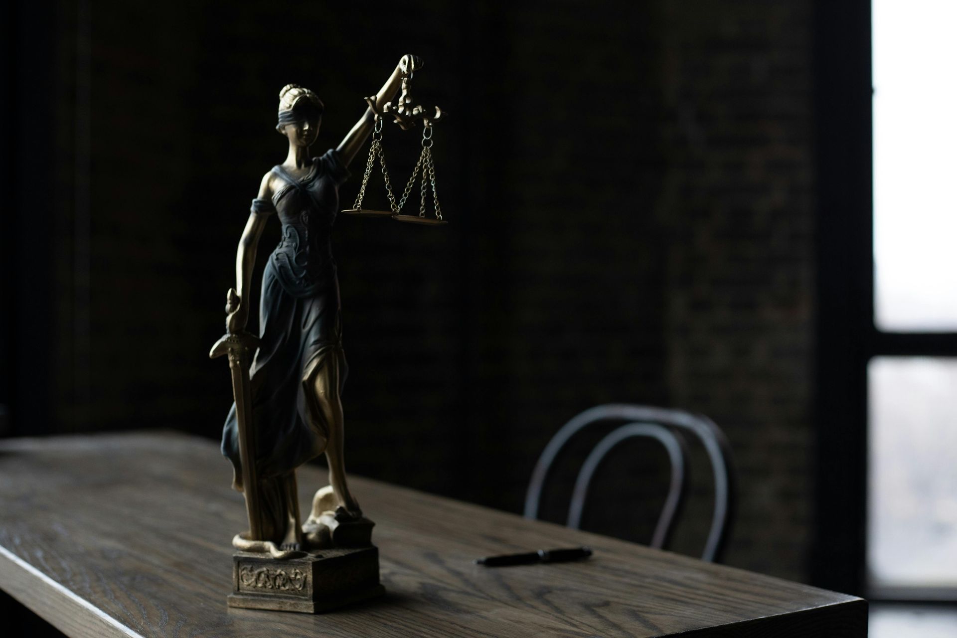 A bronze Lady Justice statue holding scales and a sword sits on a wooden desk in a dimly lit office.