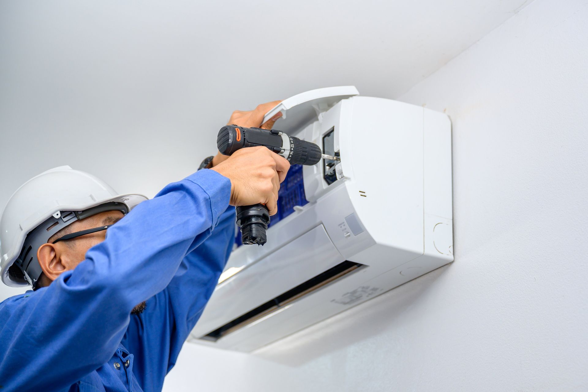 Technician in blue shirt installs a white wall-mounted air conditioner with a drill