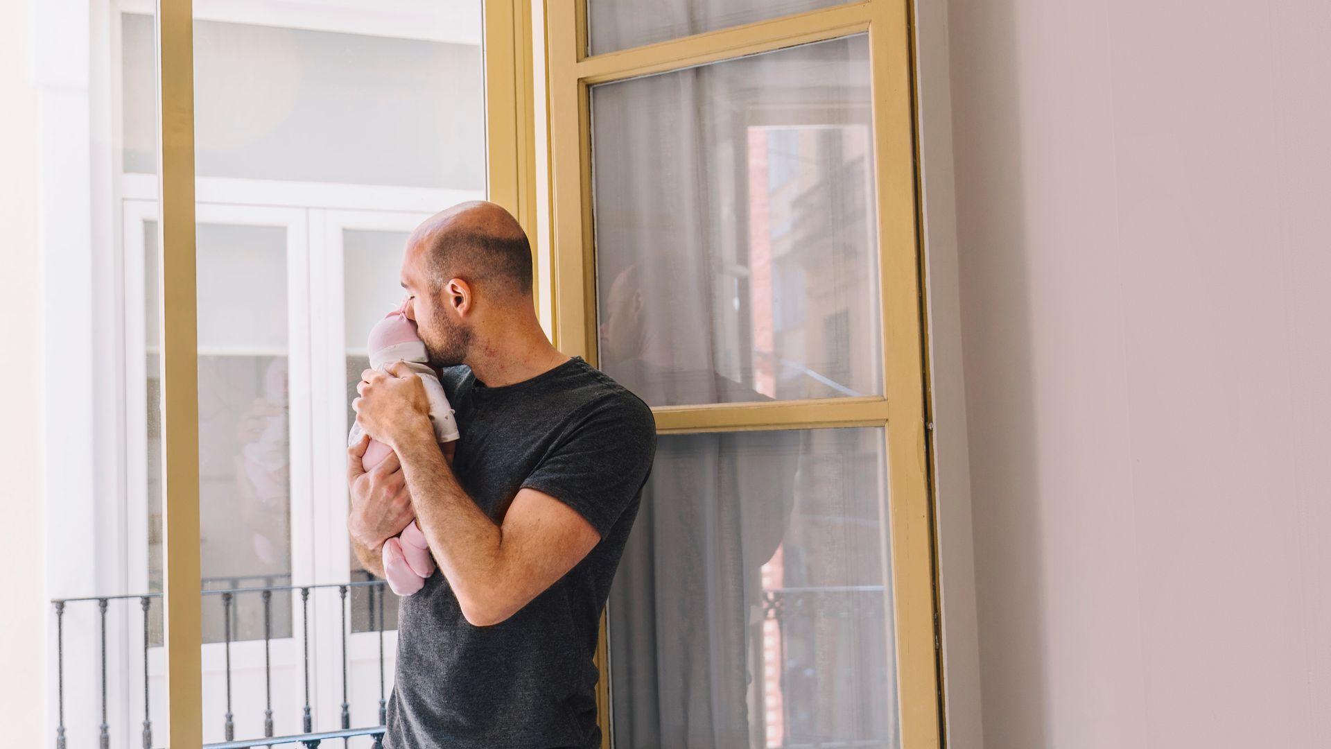 A man is holding a teddy bear and looking out of a window.