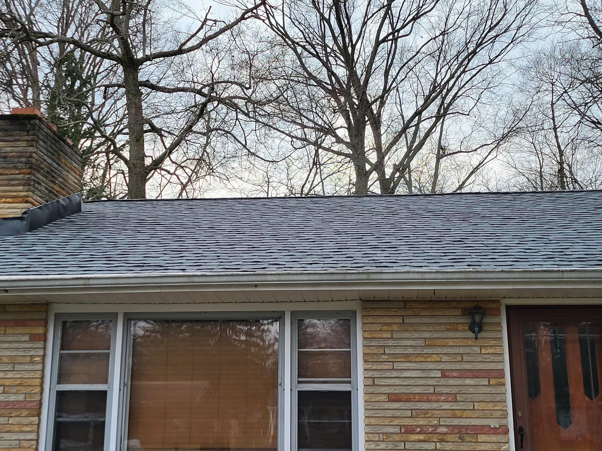 Snow-covered roof above a brick house with windows and a front door, bare trees in back