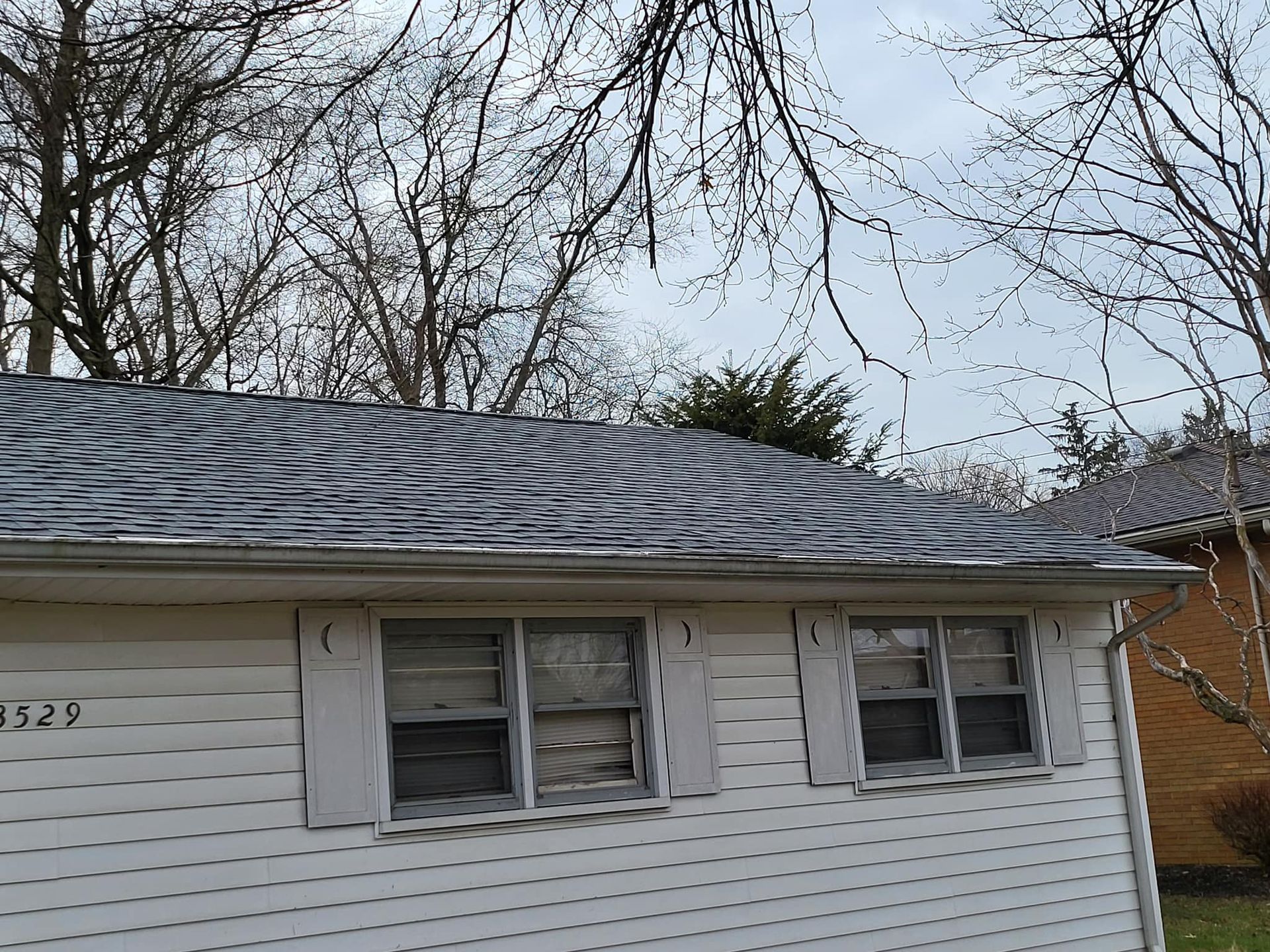 Small white house exterior with gray shingled roof and bare trees behind it