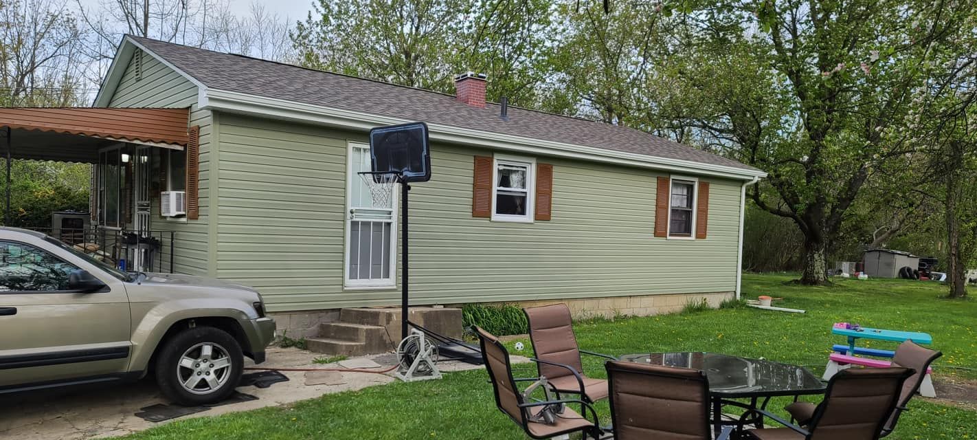 Small beige house with a parked SUV and patio chairs in a grassy yard, trees in the background