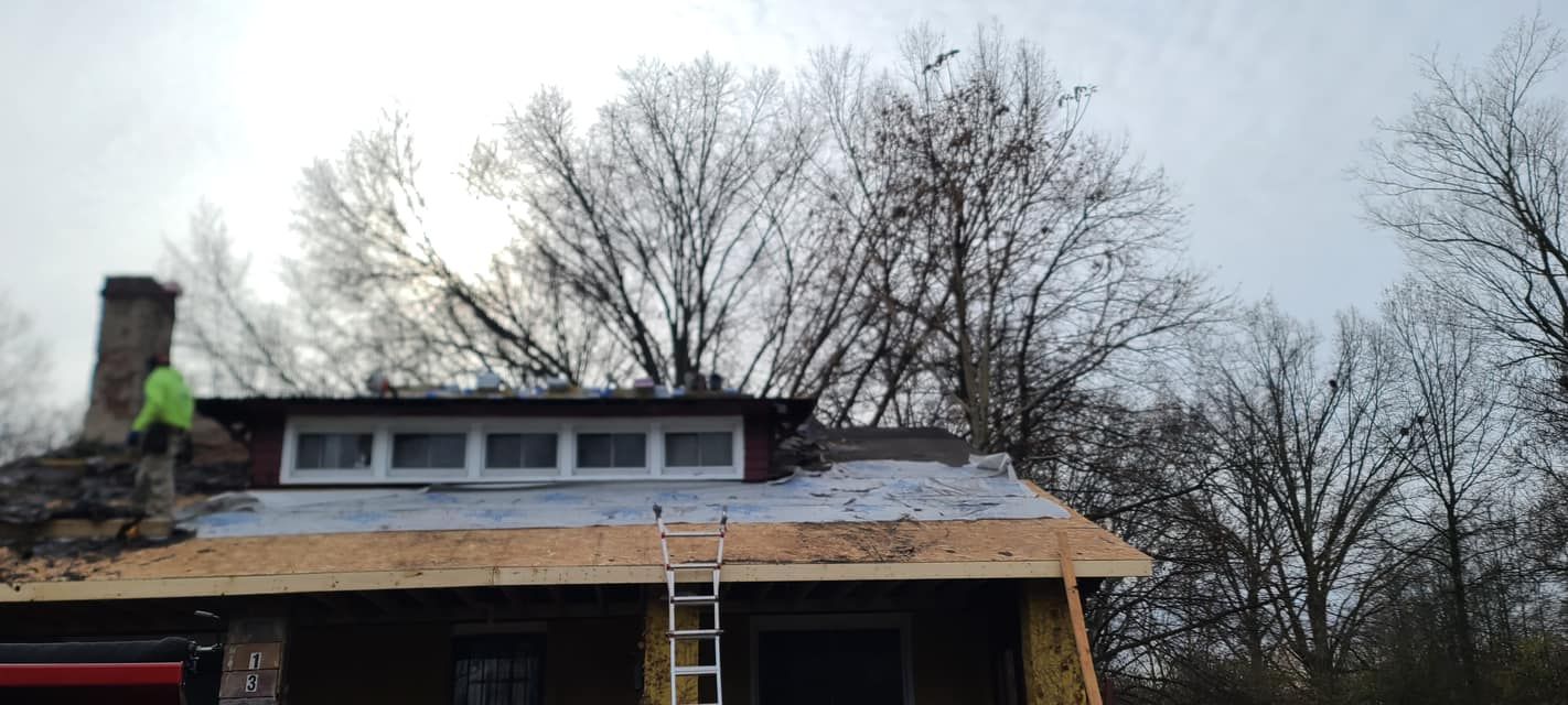Roofing work on a house with ladder, exposed framing, and cloudy winter trees behind