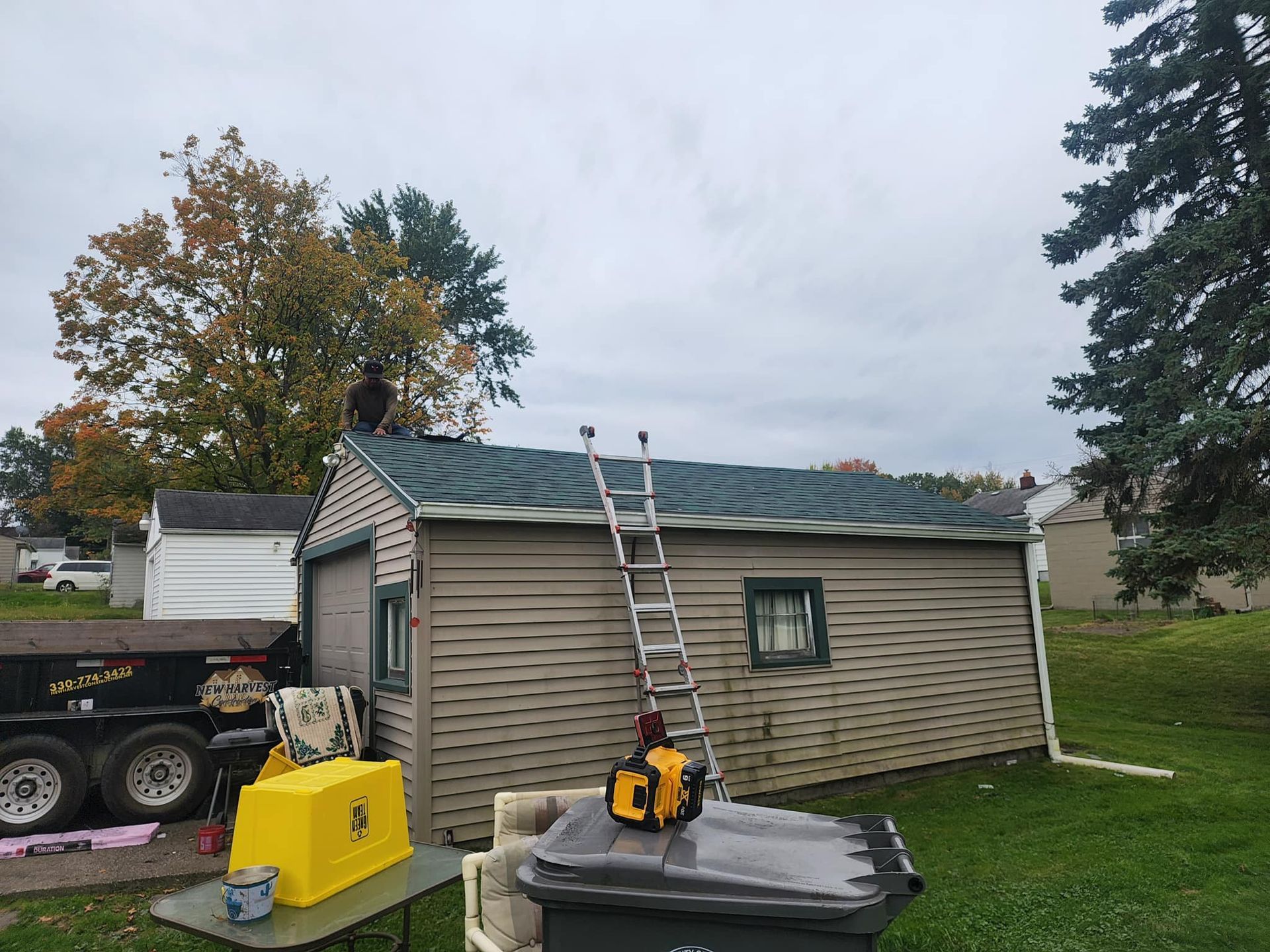 Two workers on a ladder replacing shingles on a small garage roof beside yard equipment.
